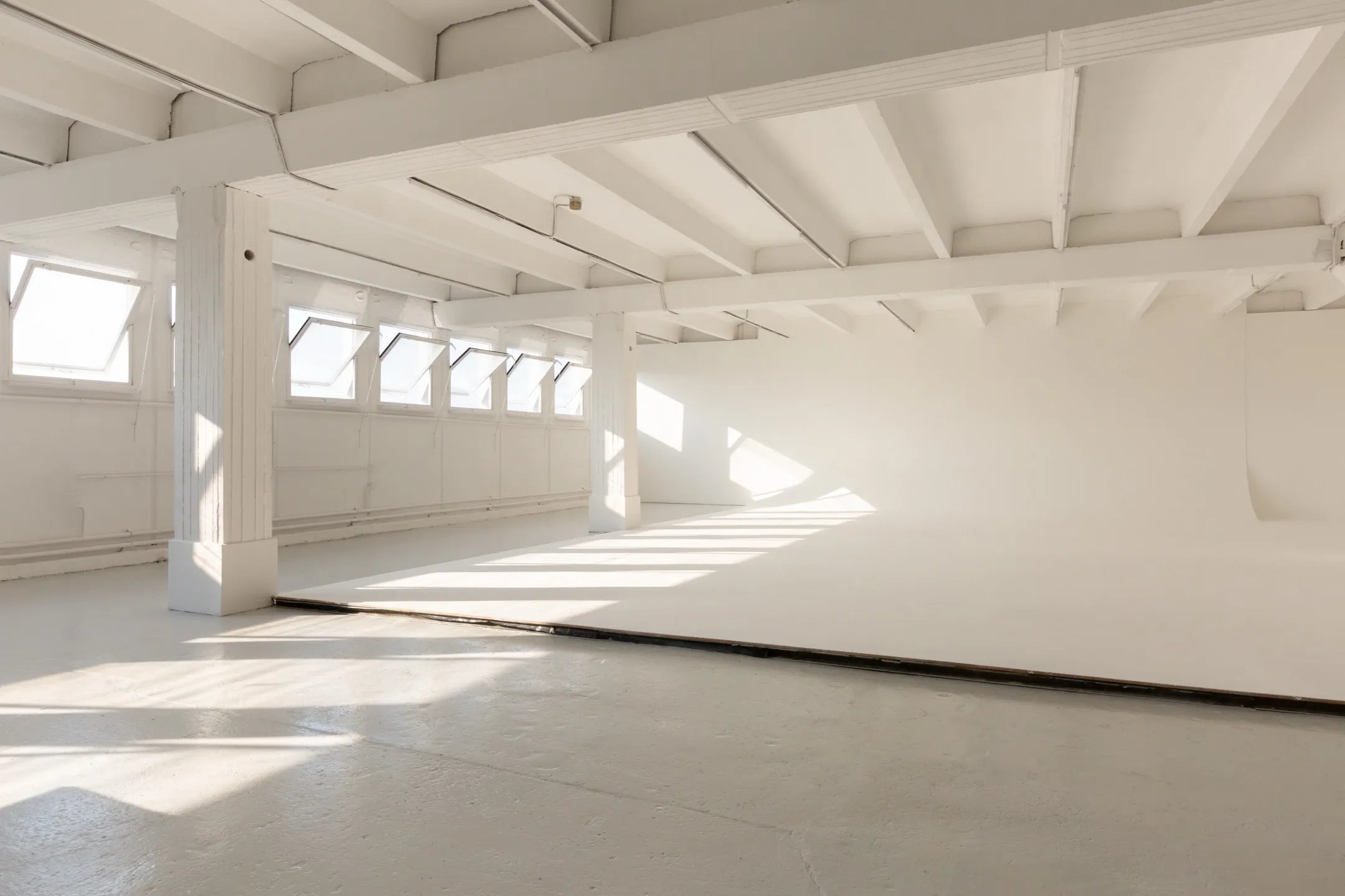 Picturepool Studios interior — bright white studio with exposed ceiling beams, a row of skylights, concrete floor and infinity cove, lit by dramatic natural sunlight